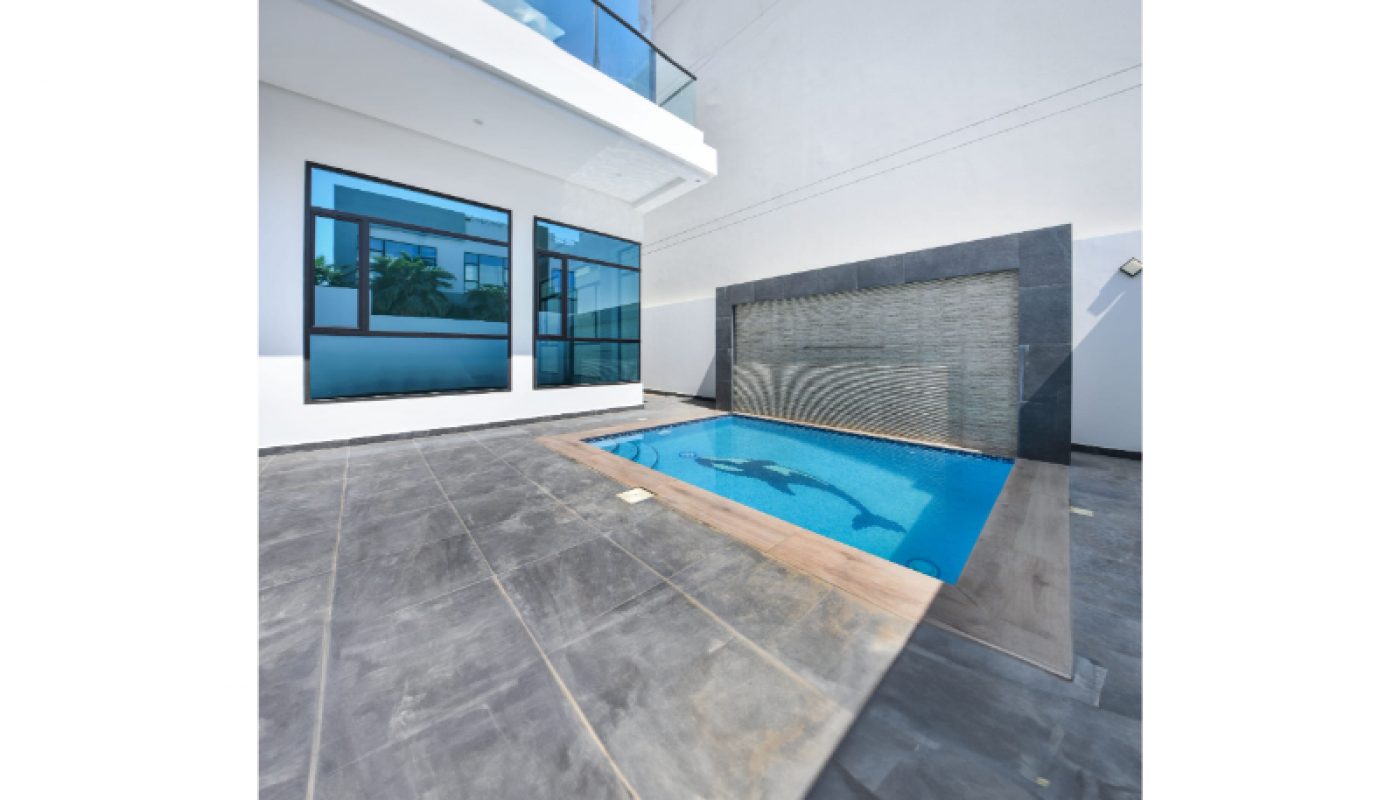 Modern swimming pool with a waterfall feature, surrounded by gray tile flooring, next to a white building with large glass windows and a balcony.