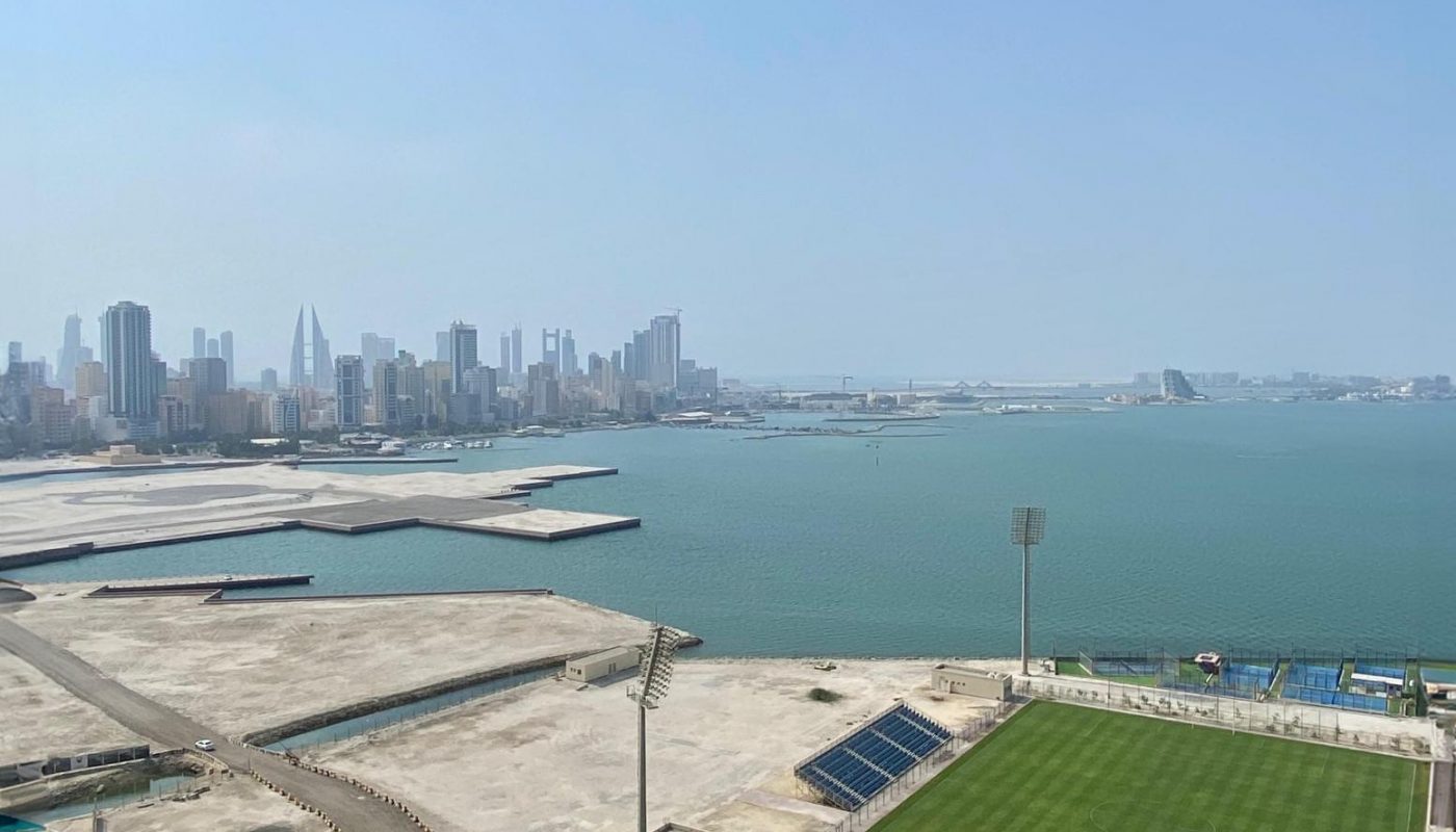 Aerial view of a coastal cityscape with a football field in the foreground, empty lots, a road, and high-rise buildings beside the sea under a clear sky.