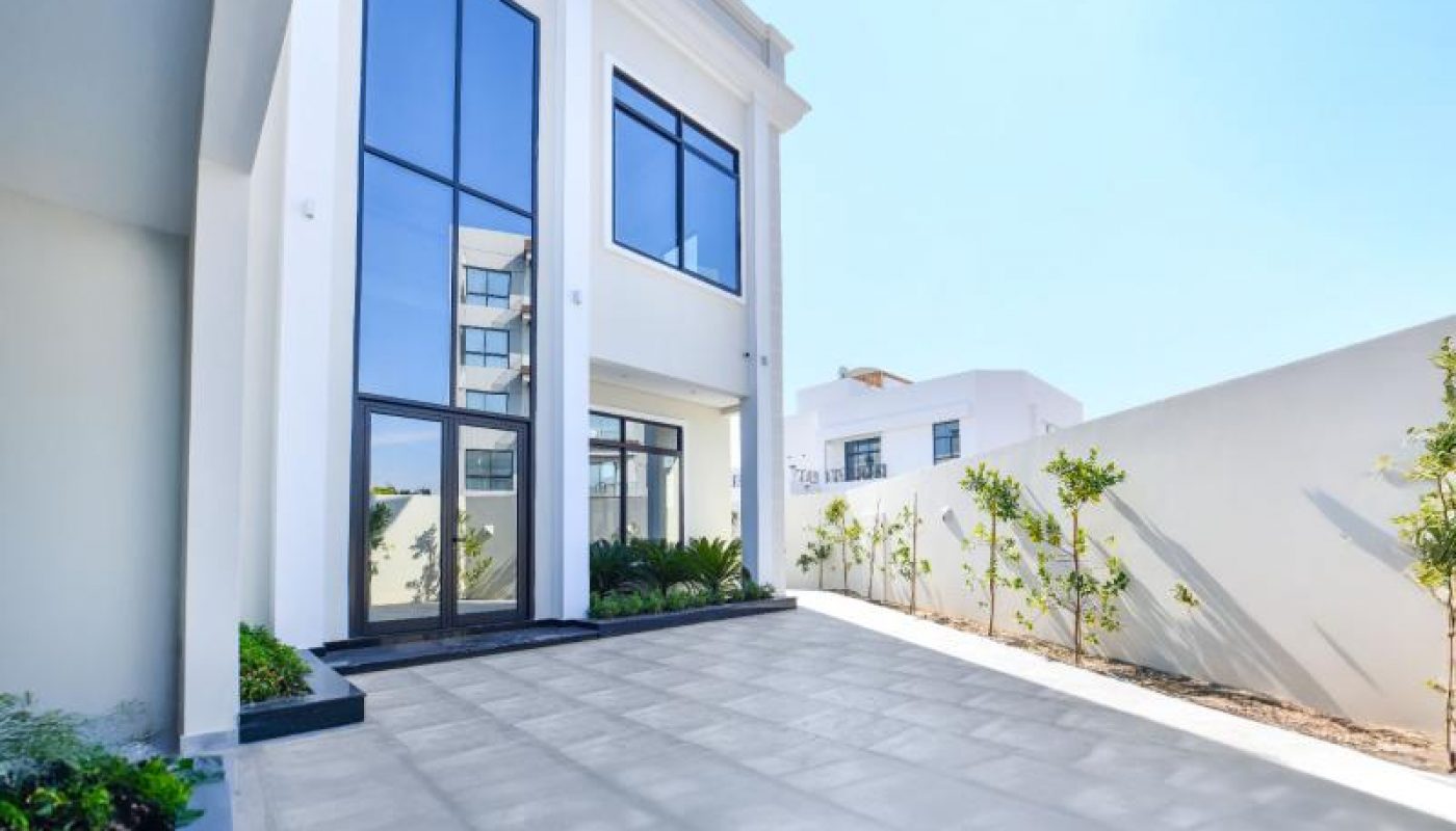 Modern two-story white house with large glass windows, a wide tiled driveway, and minimal landscaping against a clear blue sky.