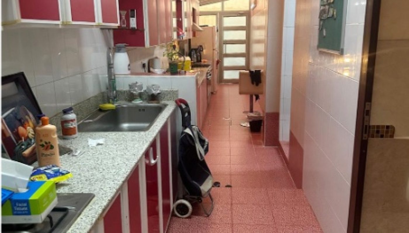 A narrow kitchen with red cabinets, white countertops, and pink patterned floor tiles. Various kitchen items are on the counter, and a hallway leads to a door at the back.