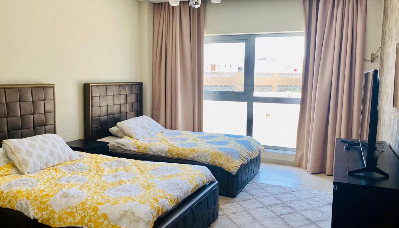 A bedroom with two single beds, each with yellow and white bedding, large window with curtains, a TV on a stand, and tiled floor.
