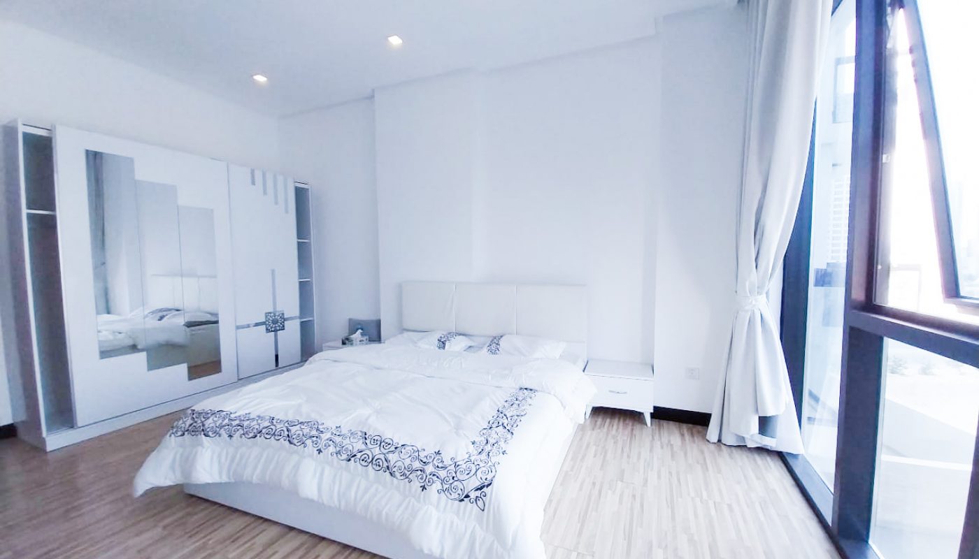 A minimalist bedroom with a large bed, white bedding, mirrored wardrobe, wooden floor, and floor-to-ceiling windows letting in natural light.