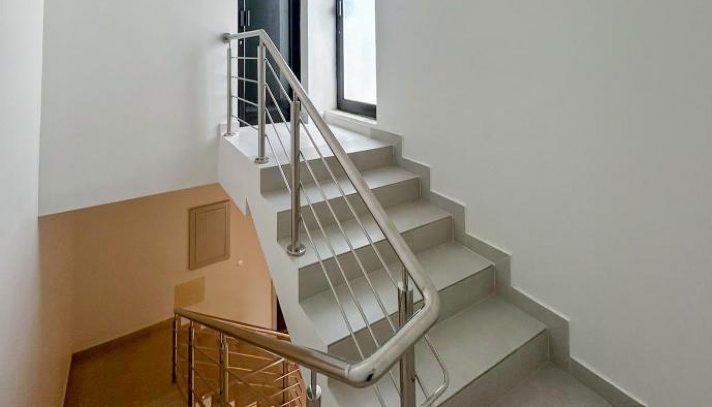 Modern indoor staircase with light grey steps, stainless steel railing, and a landing leading to a black door and a window. Walls are painted white and the area is well-lit.