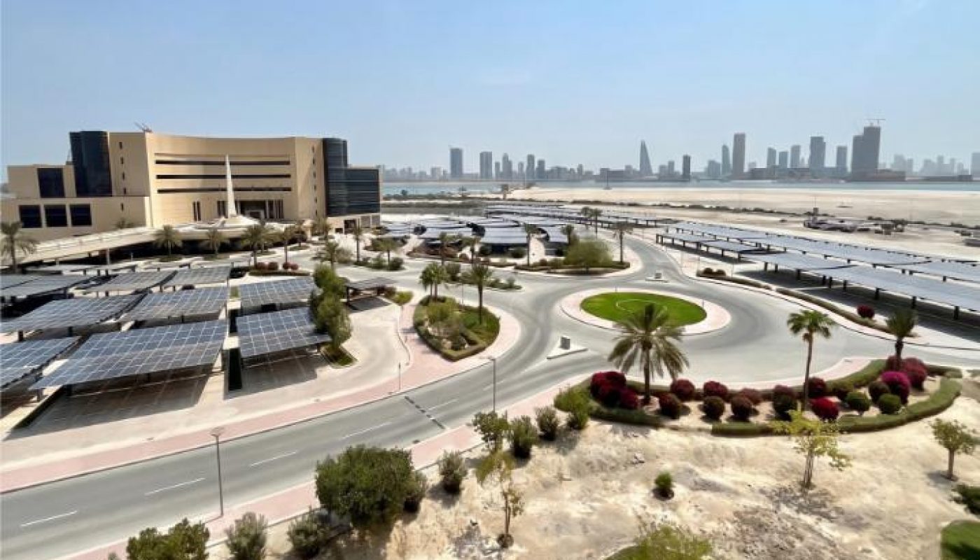A modern building with solar panel-covered parking, landscaped roundabout, and city skyline in the background under a clear blue sky.