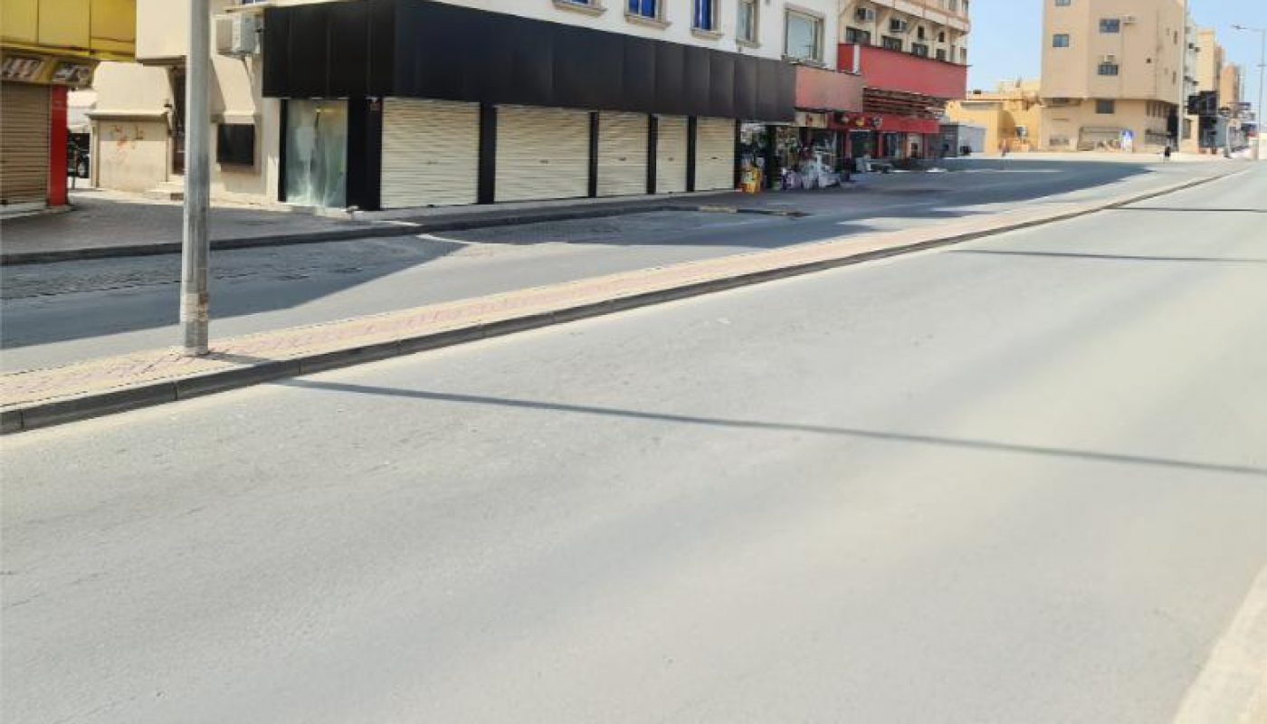 A quiet, empty street with a few closed shops in a commercial area, bordered by multi-story beige buildings under a clear blue sky.
