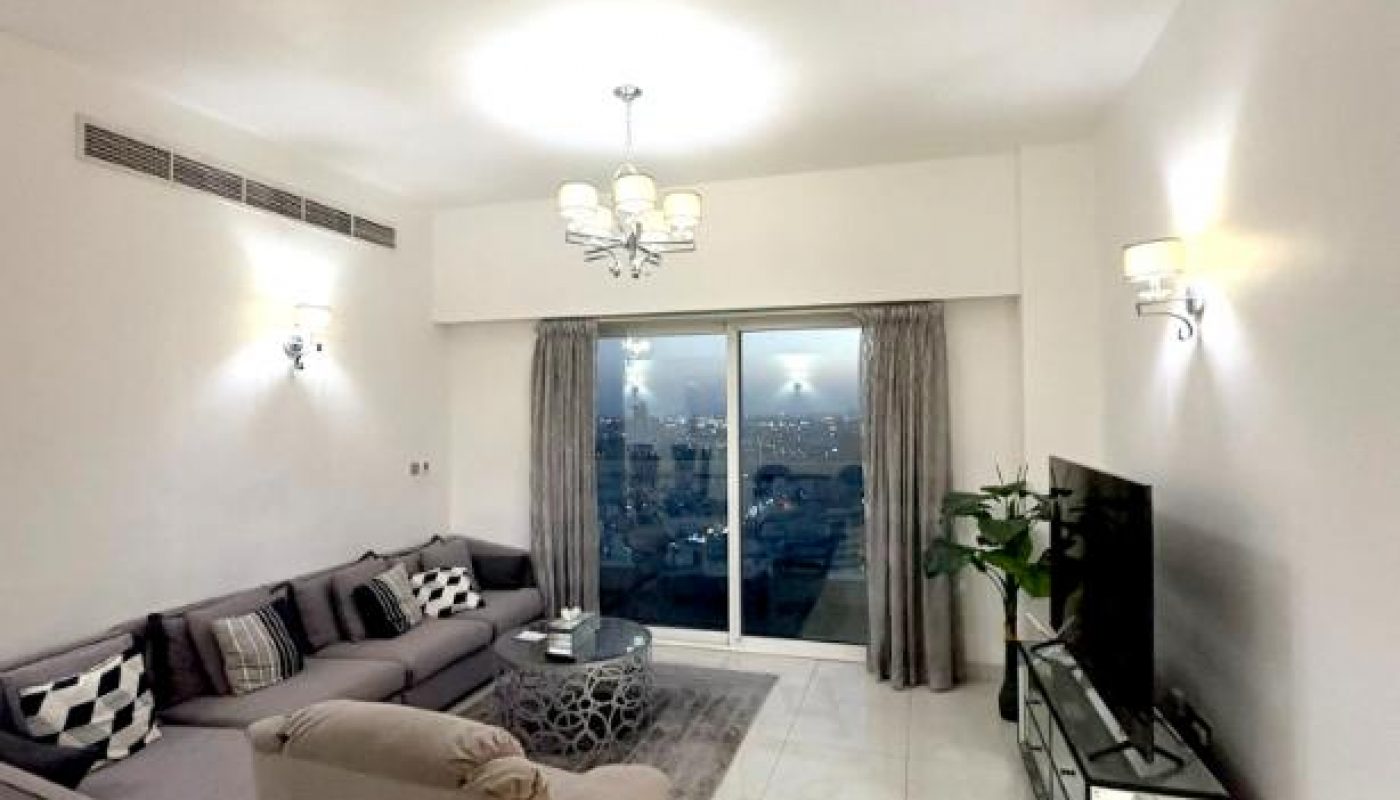 Modern living room with gray sectional sofa, armchair, TV on a stand, glass coffee table, and large window with city view. White tiled floor and neutral decor.