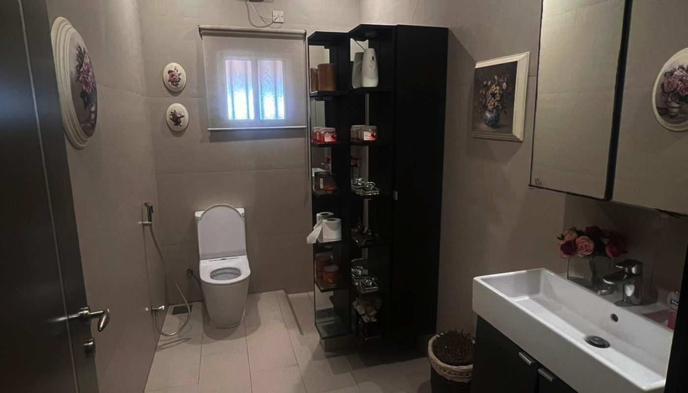 Modern bathroom with a toilet, black storage shelves, wall art, sink with mirror cabinet, and a window near the ceiling.