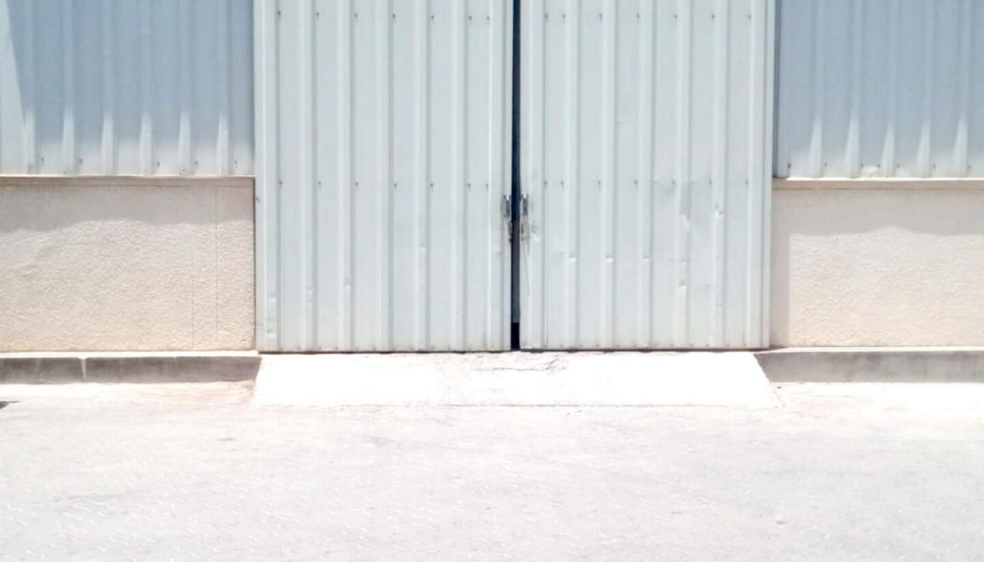 A large closed metal warehouse door with a corrugated surface, beige lower panels, and a security camera above, under a clear blue sky.