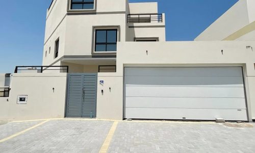 A modern, multi-story white building with large windows and a big garage door, set against a clear blue sky. A paved driveway with a yellow border leads to the entrance and garage. This elegant villa in Diyar Al Muharraq is now for sale.