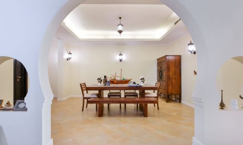 A spacious room in the 3BR Villa at Al Areen features white walls and a wooden dining table with chairs under drop lantern lights. The table, adorned with a model ship, is surrounded by art pieces set in arch-framed walls. This villa also boasts a big garden for ultimate relaxation.