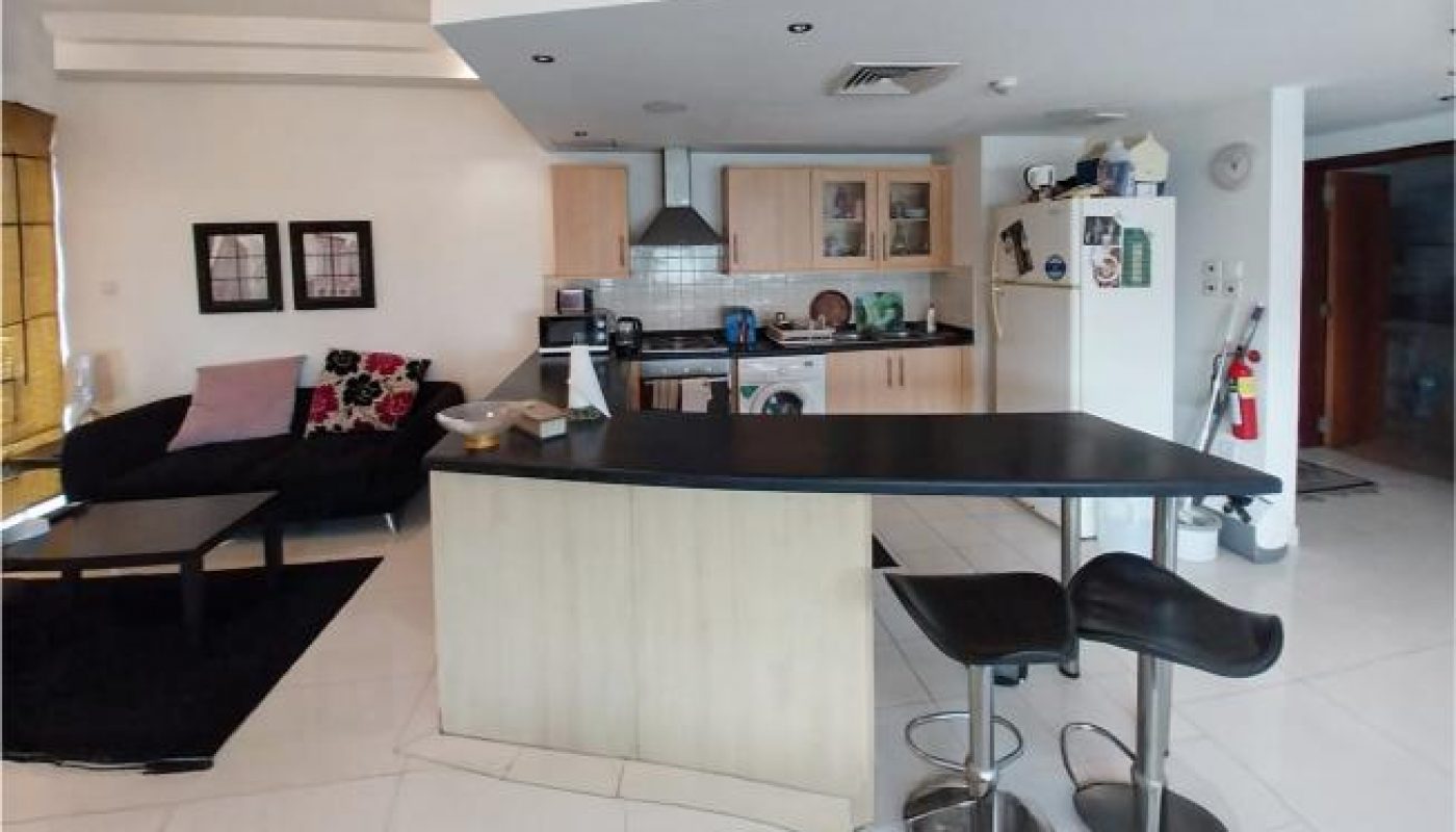 Modern open-plan kitchen and living area with black countertops, bar stools, light wood cabinets, and a black sofa with patterned cushions.