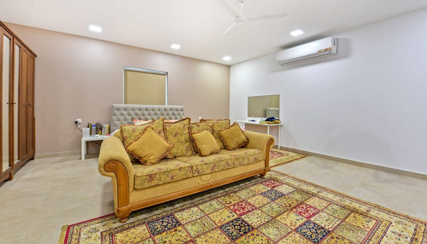 Spacious room with a large patterned rug, a mustard sofa with cushions, air conditioner, ceiling fan, and a dresser. A window and a table with decorative items are seen.