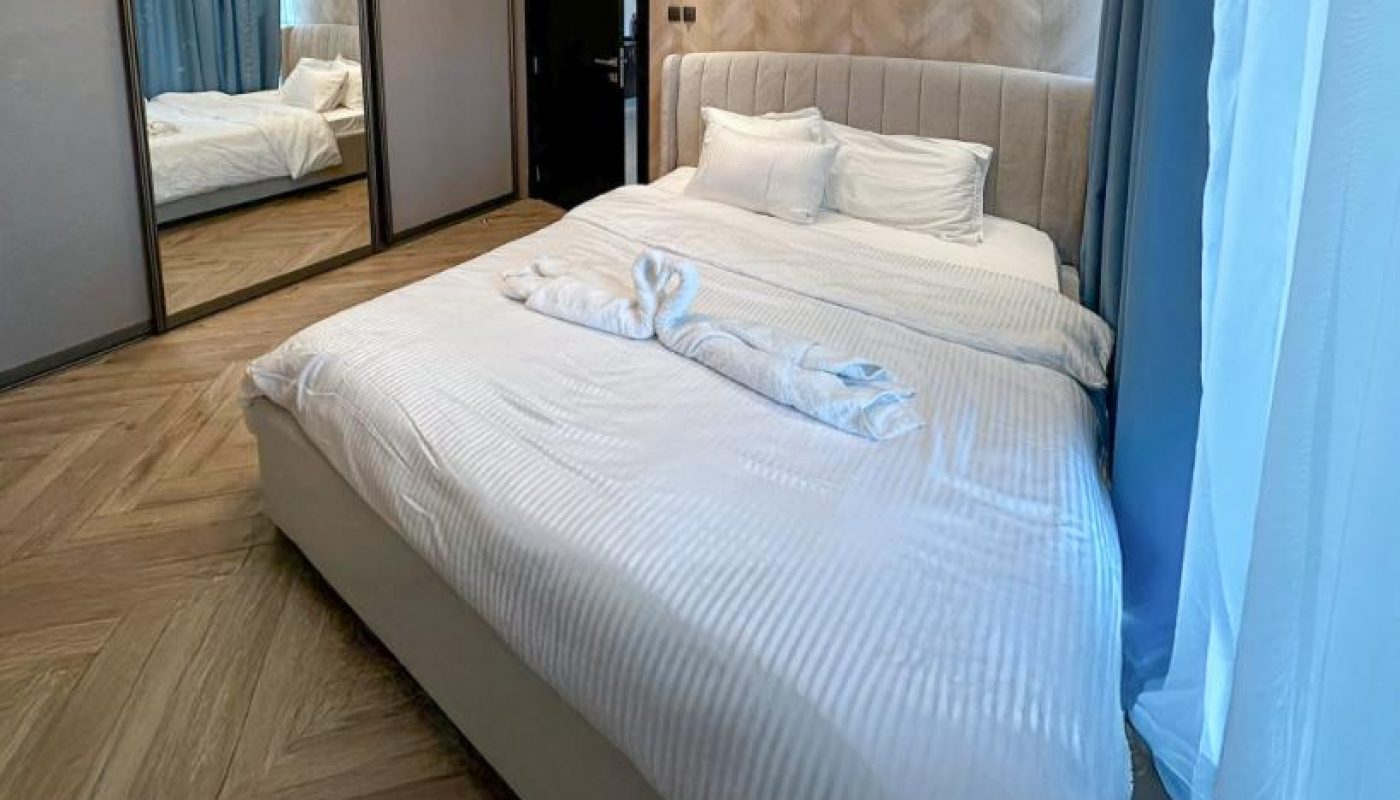 Modern bedroom with a large bed, white bedding, a towel folded into a swan shape, mirrored closet doors, wooden herringbone floor, and a window with sheer curtains.
