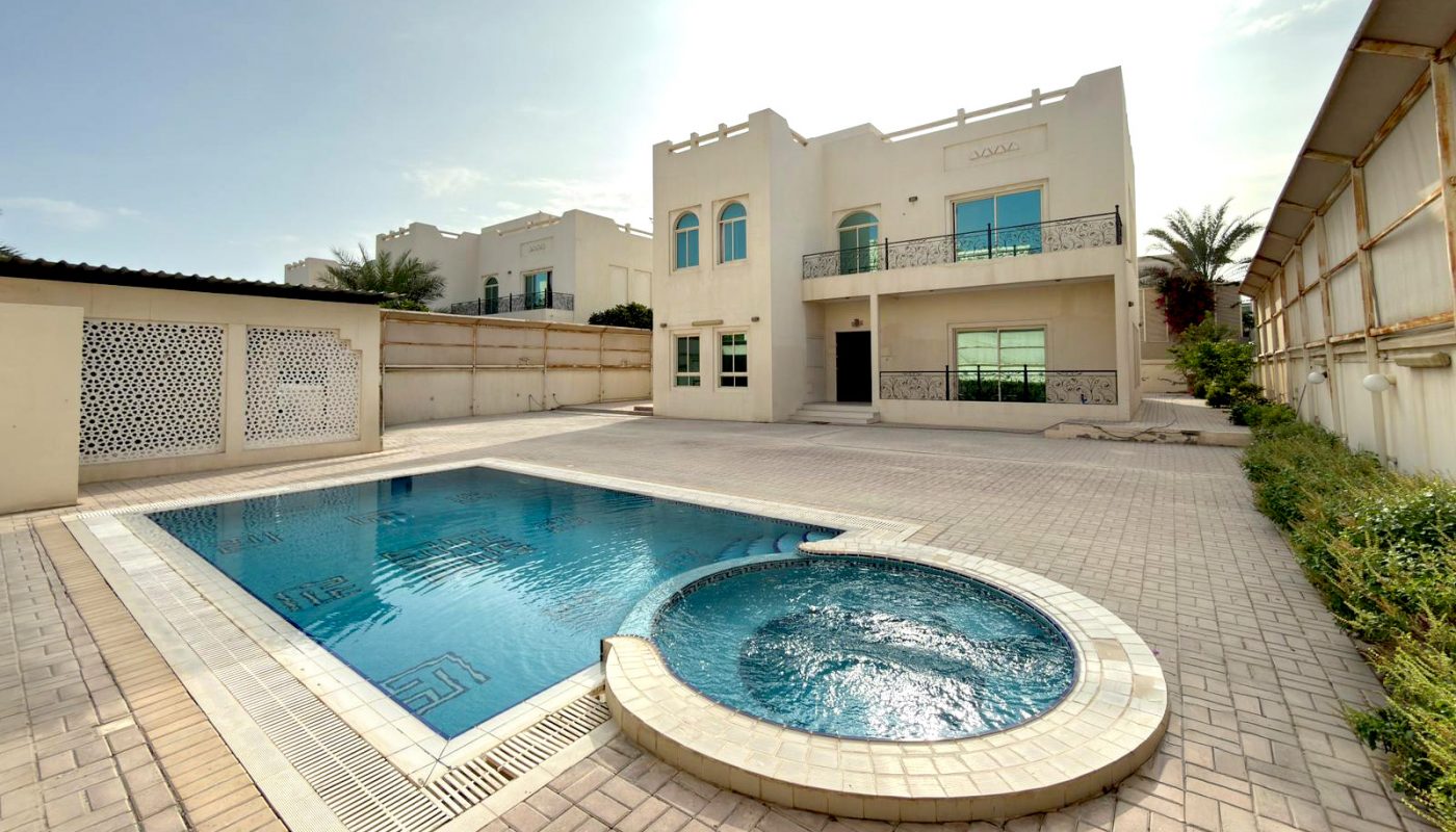 A modern two-story house with a large, paved backyard featuring a rectangular swimming pool and a round hot tub under a partly cloudy sky.
