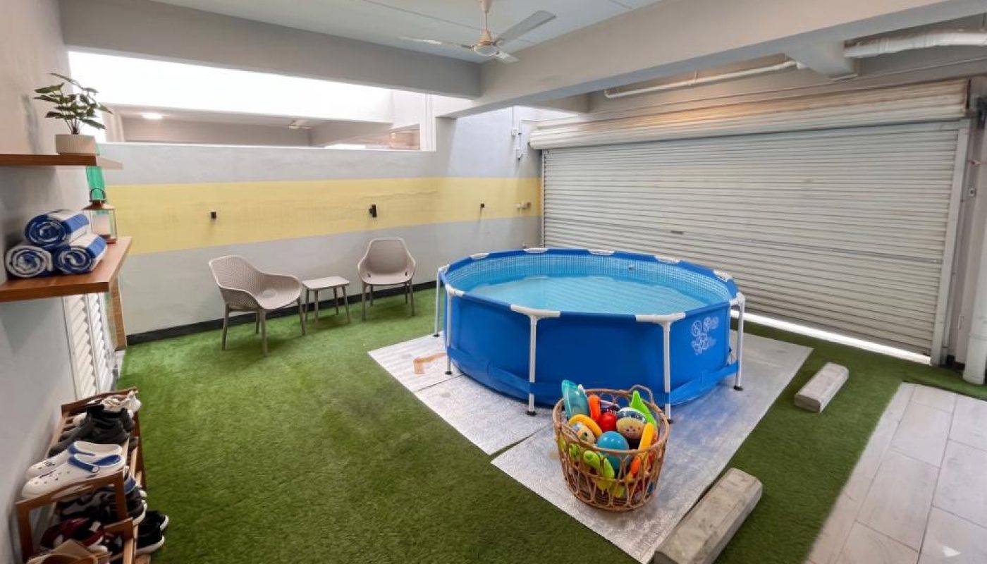 An above-ground pool with toys sits on artificial grass in a garage-like space with a roll-up door.