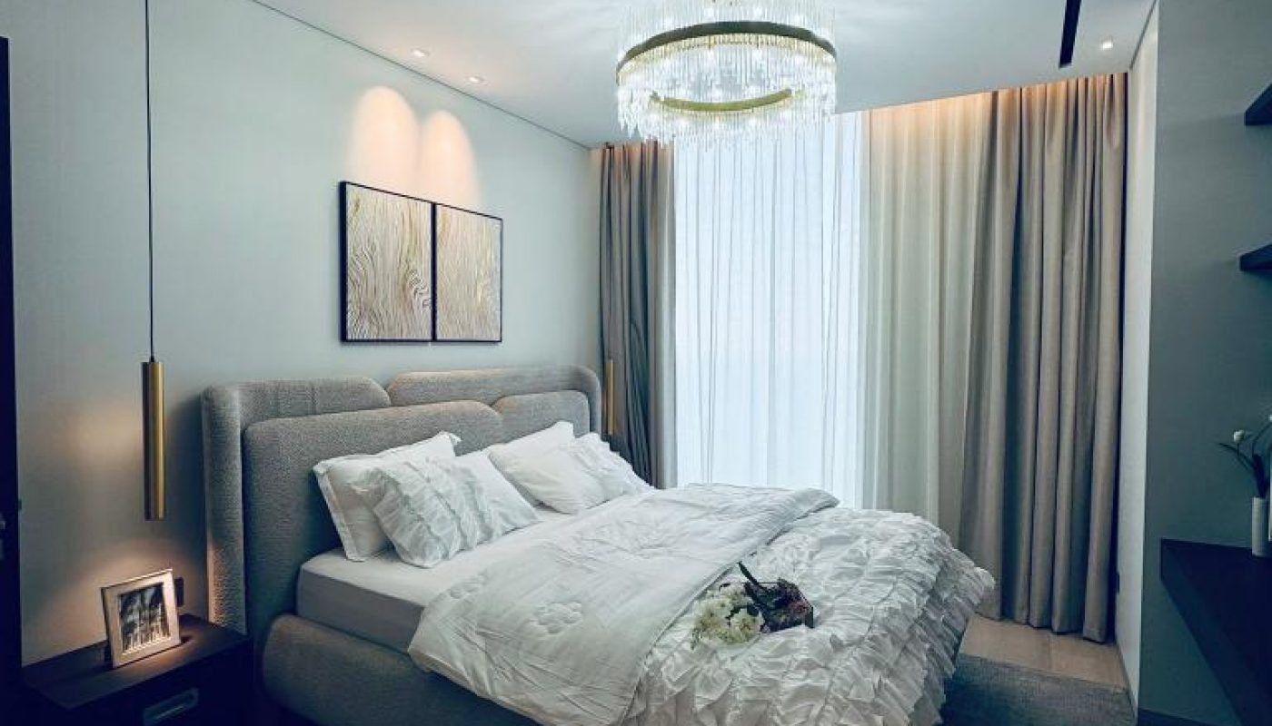 A modern bedroom with a large bed, white bedding, neutral walls, two framed artworks, a chandelier, and floor-to-ceiling curtains letting in natural light.