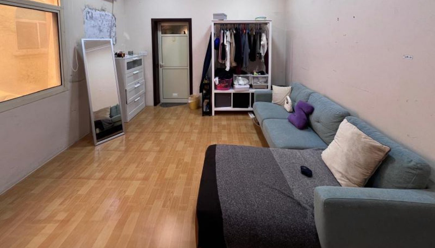 A tidy bedroom with wooden flooring, a blue sofa bed with cushions and a blanket, an open wardrobe, a standing mirror, a dresser, and an air conditioner on the wall.