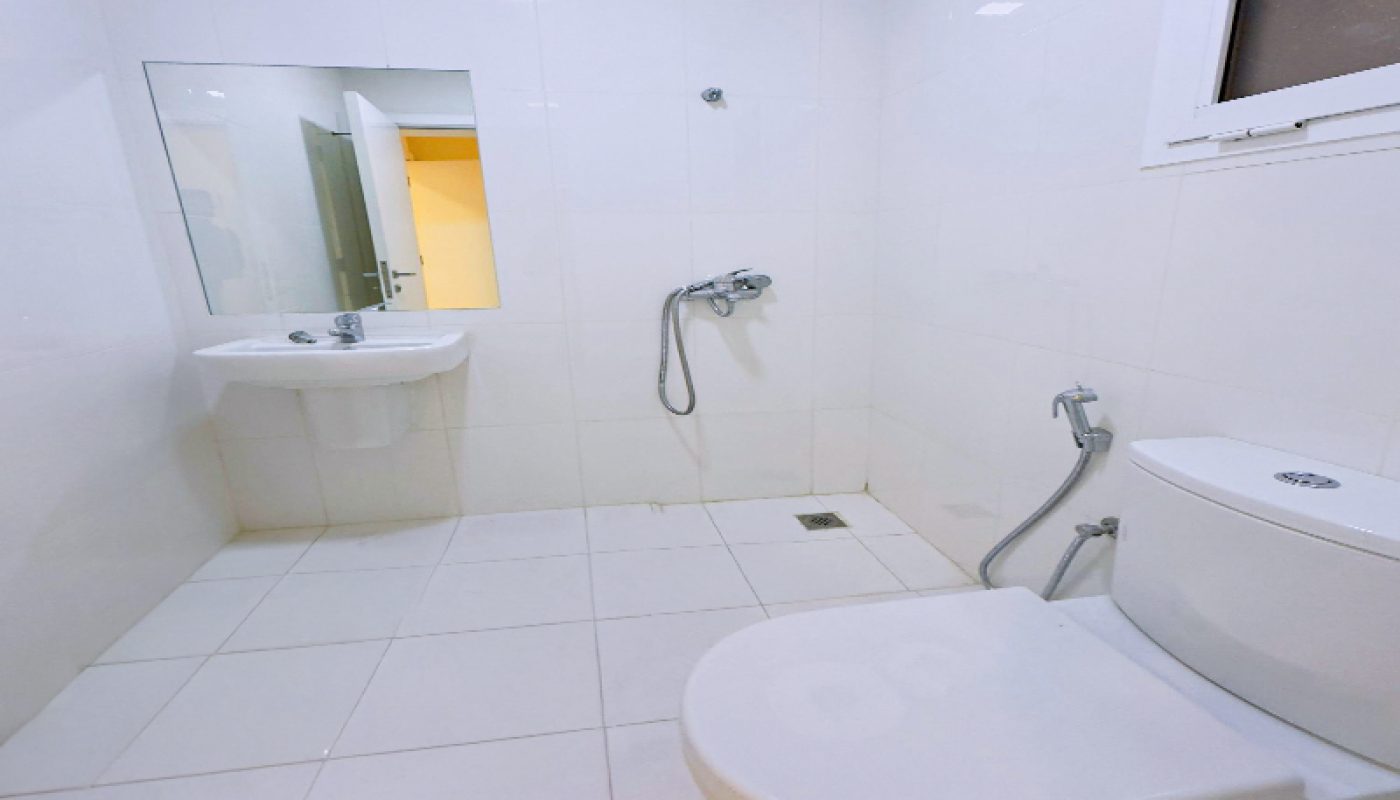 A clean, white-tiled bathroom with a toilet, wall-mounted sink, mirror, handheld showerhead, and a window.