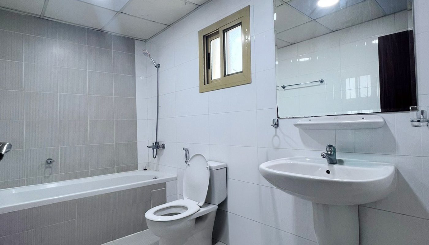 Modern bathroom with bathtub, toilet, wall-mounted sink, large mirror, tiled walls and floor, ceiling lights, and a small window.