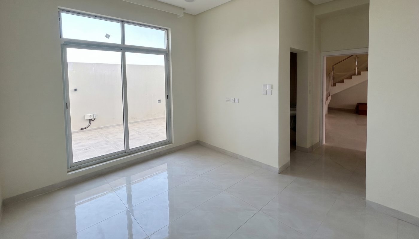 Unfurnished empty room with large tiled floor, white walls, ceiling fan, and a window overlooking an outdoor area; open doorway leads to hallway with stairs.