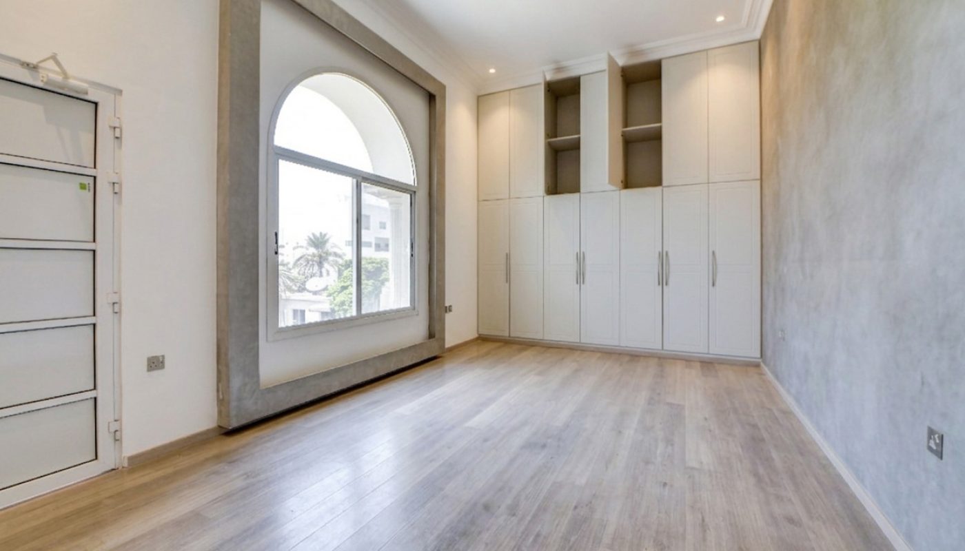 Empty room with wood flooring, large arched window, built-in white cabinets, and light grey walls. Outdoor greenery is visible through the window.