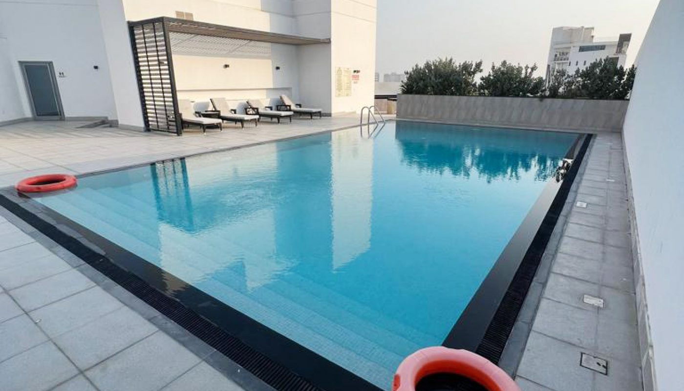 Rooftop swimming pool with clear blue water, surrounded by white tiles and orange lifebuoys, with lounge chairs and a modern building in the background.