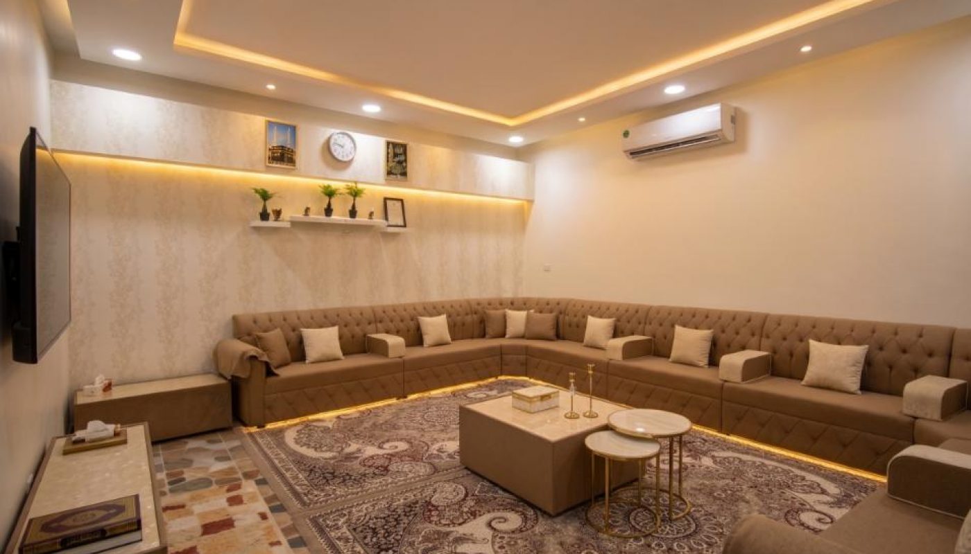 A spacious living room with U-shaped brown sofas, patterned rugs, and wall shelves holding decor items. The room features recessed lighting and a wall-mounted air conditioner.