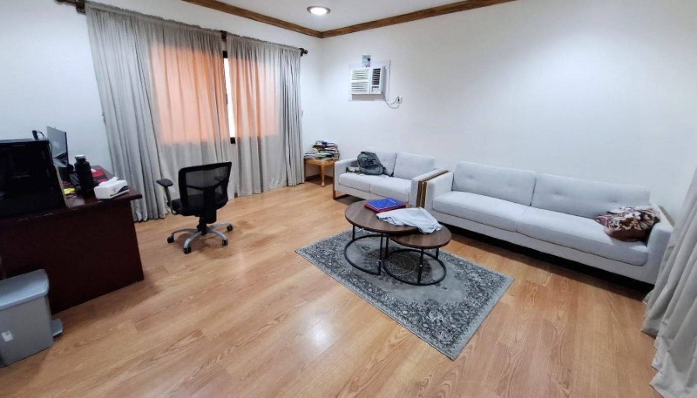 A tidy room with wood flooring, a desk and chair, a sofa, a small rug with a coffee table, and an air conditioner on the wall.