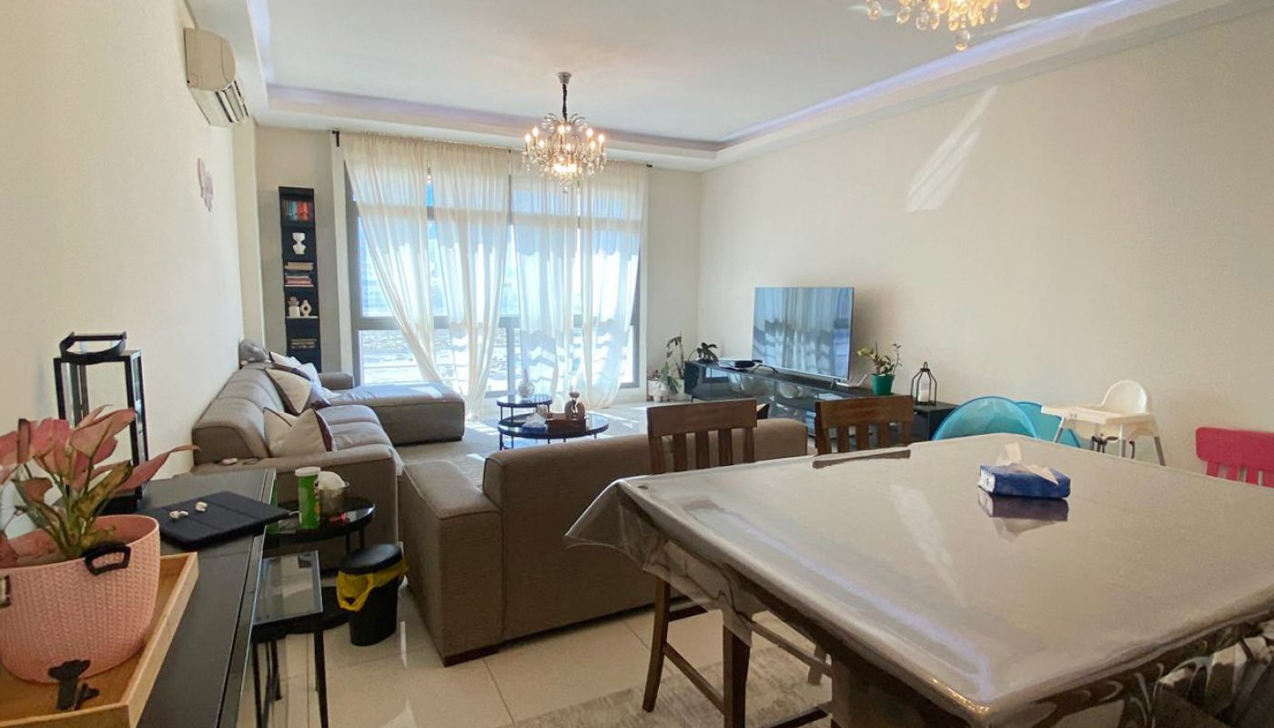A living and dining room with a beige sofa, dining table, TV, large window with sheer curtains, and two chandeliers.
