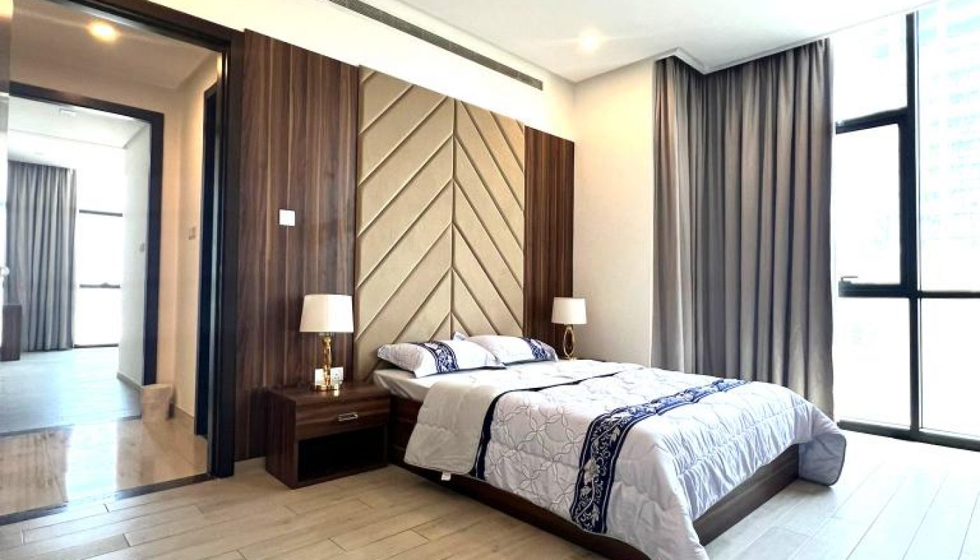 A modern bedroom with a double bed, wooden accent wall, two bedside tables with lamps, large windows with gray curtains, and light wood flooring.