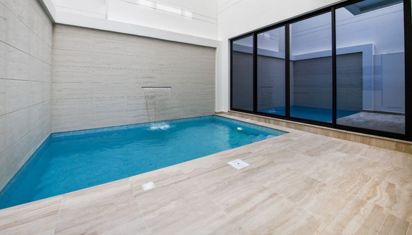 Indoor swimming pool with a waterfall feature, surrounded by beige stone tiles, and large glass windows.