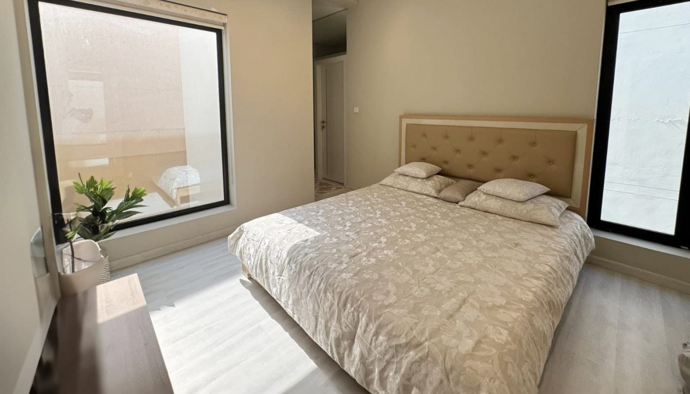 Modern bedroom with a large bed, beige headboard, two windows, air conditioner, a potted plant, and a wooden dresser. Walls and floor are light-colored.