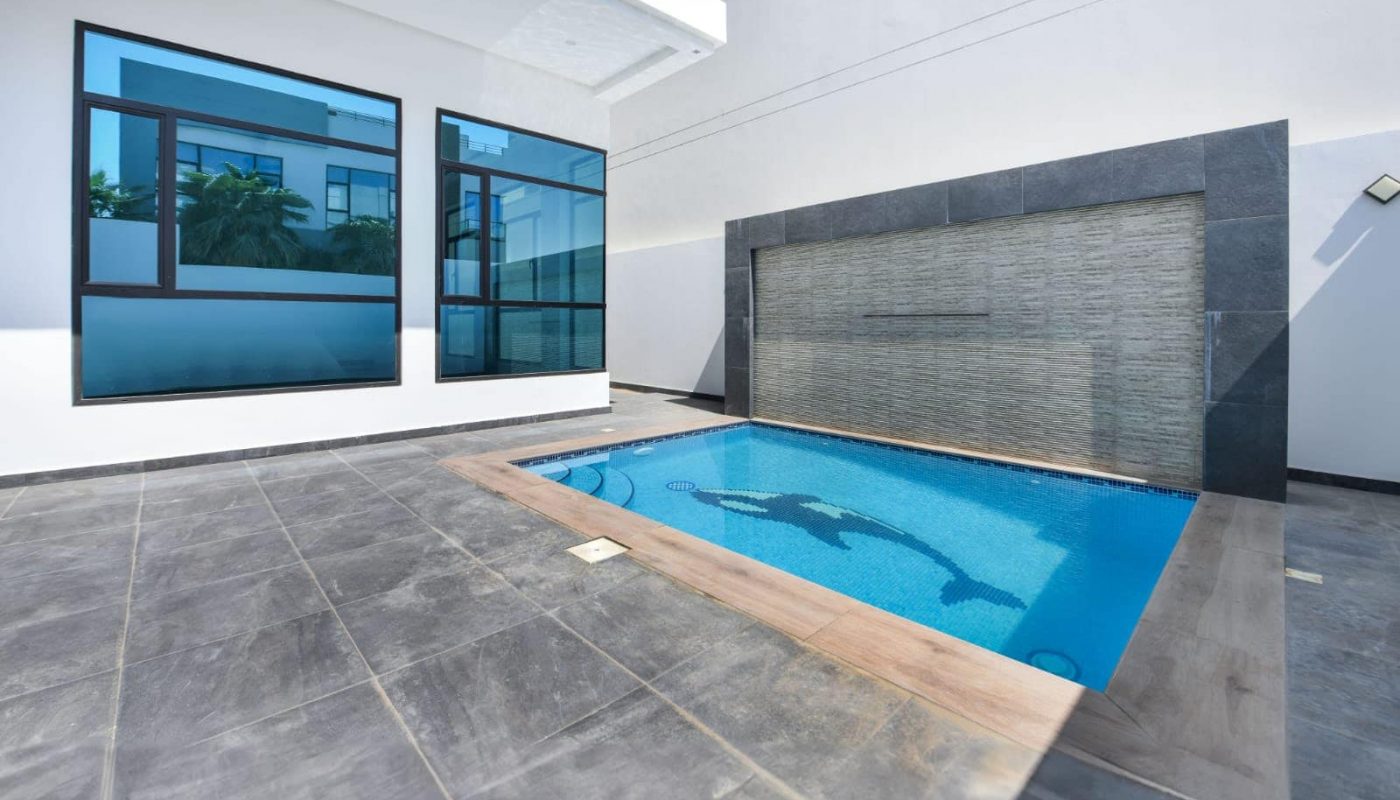 Modern courtyard with a small rectangular pool, surrounded by a tall building, glass windows, and a stone-tiled floor.