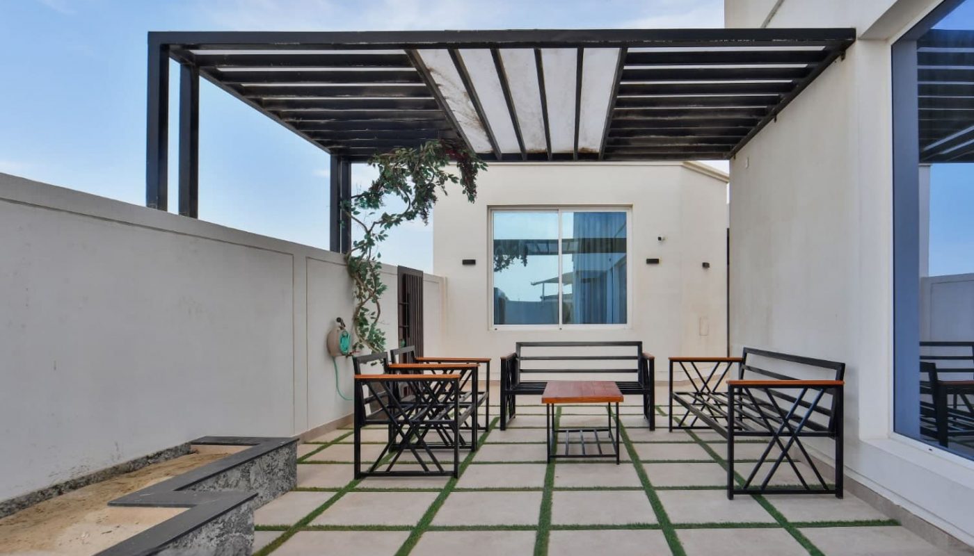Modern patio with metal pergola, two benches, and small tables on a tiled floor with grass strips. White building walls and a clear blue sky are visible in the background.