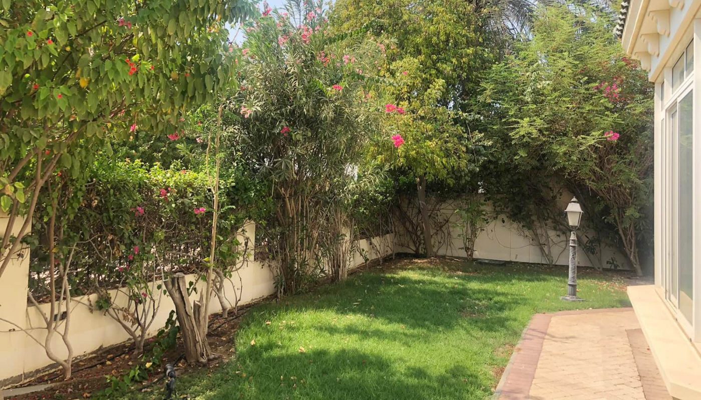 A spacious garden with green grass, various trees, and flowering plants next to a beige 4BR villa in Hamala. A paved walkway runs alongside the garden, and there is a street lamp in the corner.