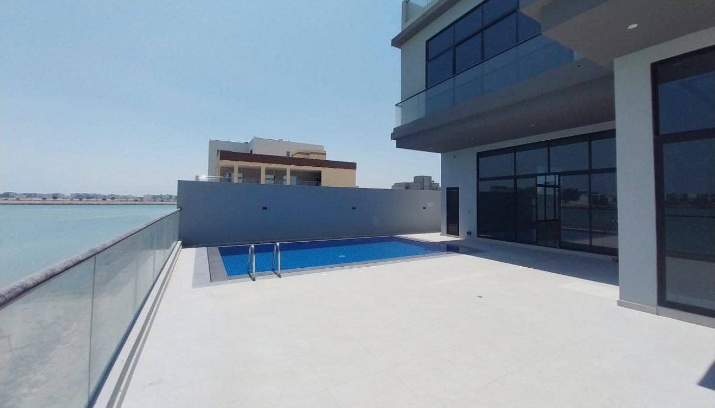 Modern waterfront building with large balconies, glass railings, and a rectangular swimming pool on a spacious tiled patio overlooking the water under a clear sky.