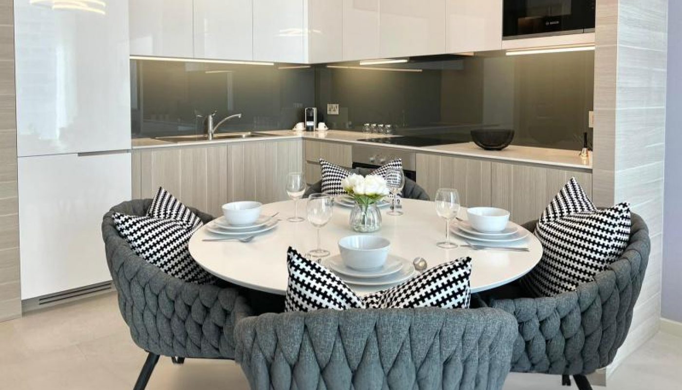Modern kitchen and dining area with a round table set for four, gray quilted chairs, and monochrome cushions, featuring sleek white cabinets and a contemporary light fixture.