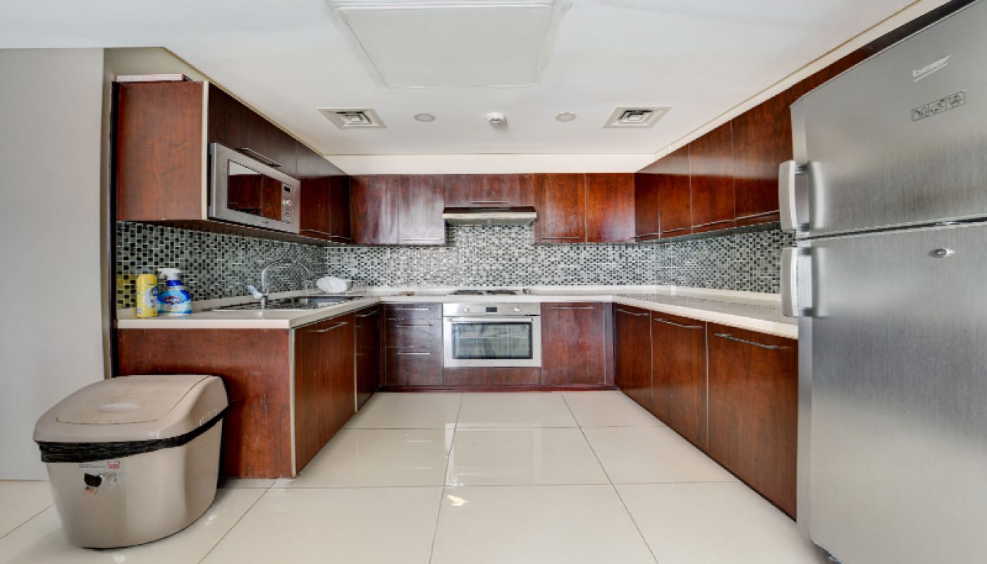Modern kitchen with dark wood cabinets, stainless steel appliances, a tiled backsplash, and a large silver trash bin on glossy white floor tiles.