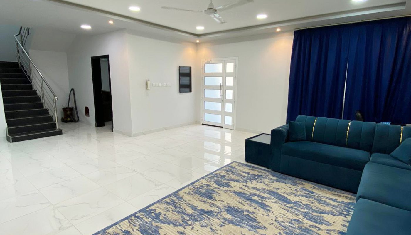Modern living room with a blue sectional sofa, blue patterned rug, white tile floor, ceiling fan, dark blue curtains, and a staircase leading upstairs.