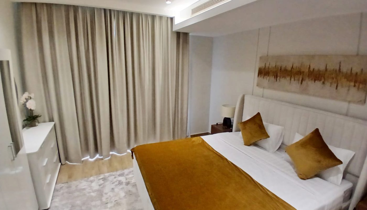 A modern bedroom in Amwaj Islands with a double bed featuring gold bedding, two gold pillows, a white headboard, a dresser, a nightstand, beige curtains, and wall art—perfect for those seeking stylish Freehold Apartments.