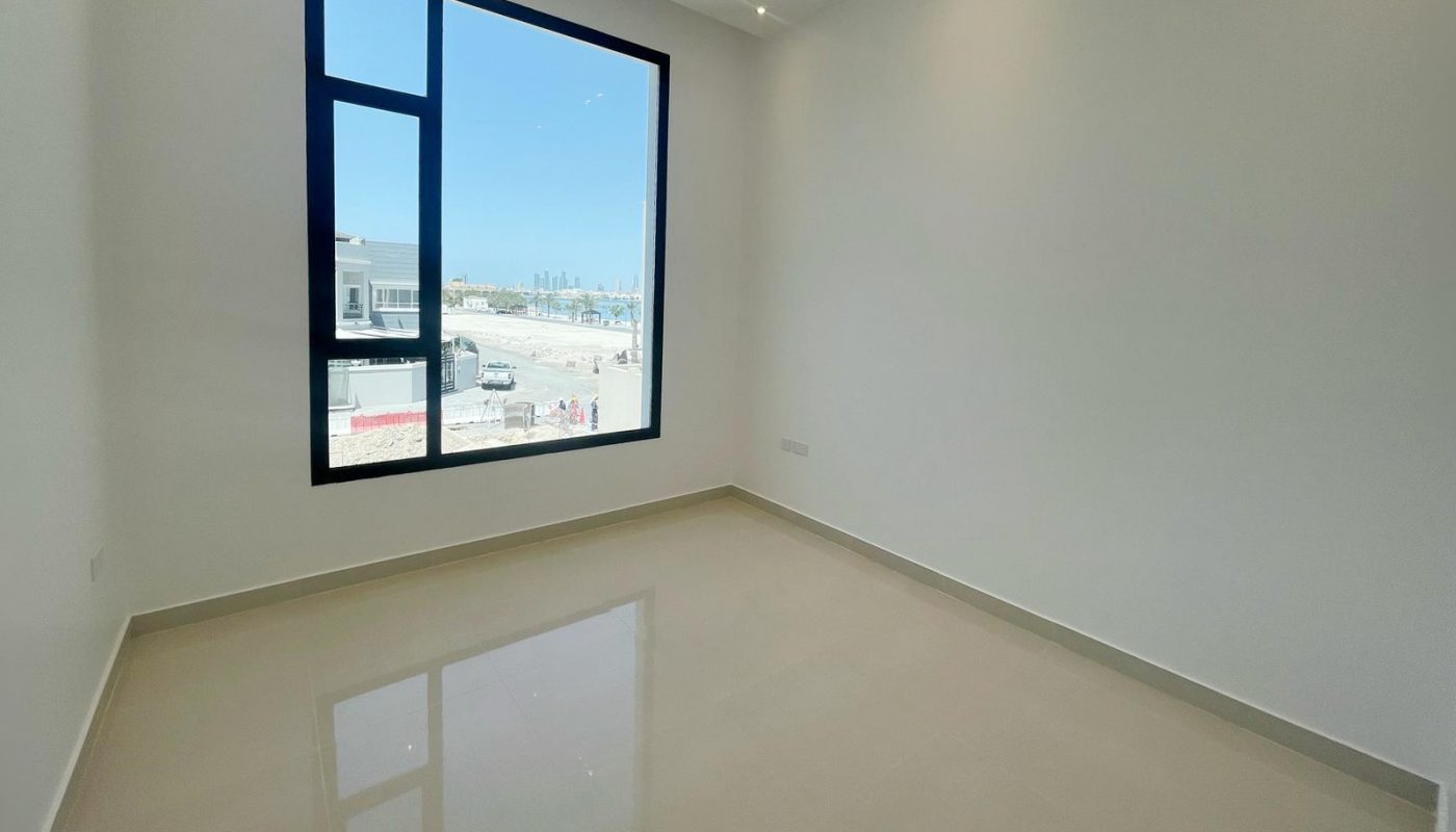 Empty room with beige tile floor, white walls, ceiling lights, and a large window overlooking a bright outdoor area with buildings and blue sky.