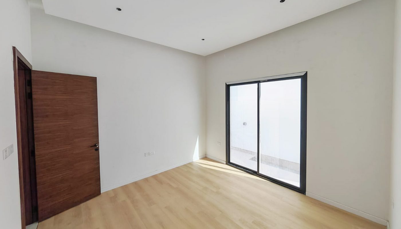 Empty, unfurnished room with light wood flooring, white walls, a wooden door on the left, and large glass sliding doors on the right letting in natural light.