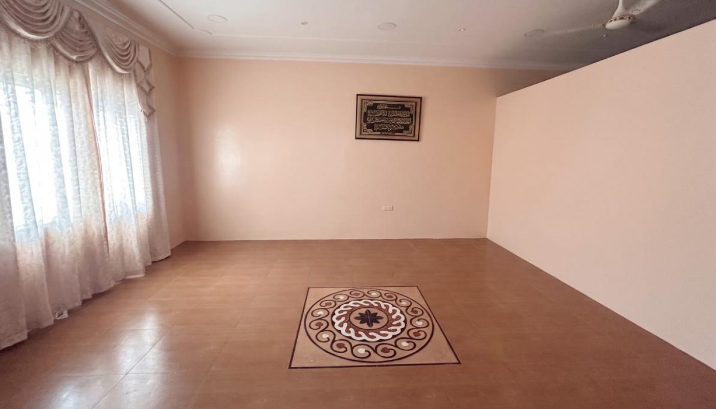A spacious, empty room with tiled flooring, a decorative circular design in the center, large windows with curtains, and a wall-mounted framed artwork.