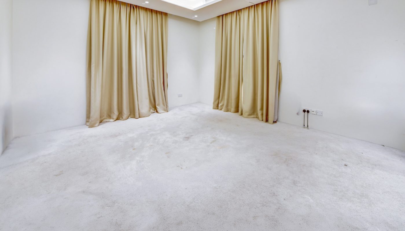Empty room with white walls, white carpet, recessed ceiling lighting, and two windows covered by beige curtains. No furniture or decor is present.