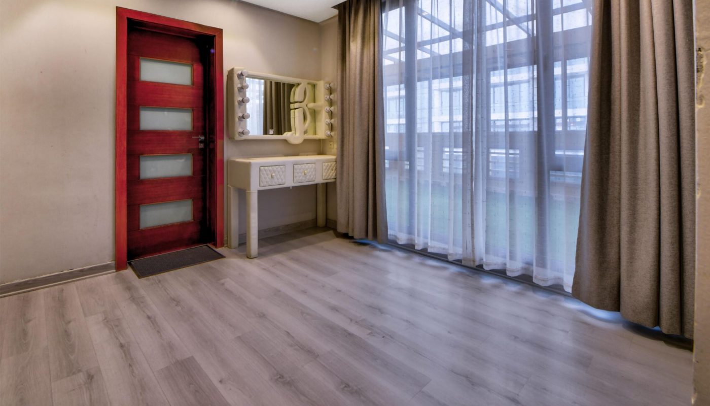 Spacious room with light wood flooring, a red door, a vanity table with a mirror, beige walls, and large windows with sheer curtains letting in natural light.