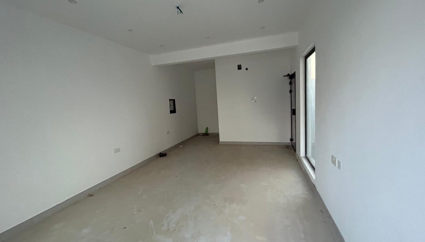 Empty, unfurnished room with white walls, bare concrete floor, recessed ceiling lights, and a large window on the right wall. There are visible electrical outlets and an unfinished ceiling fixture.