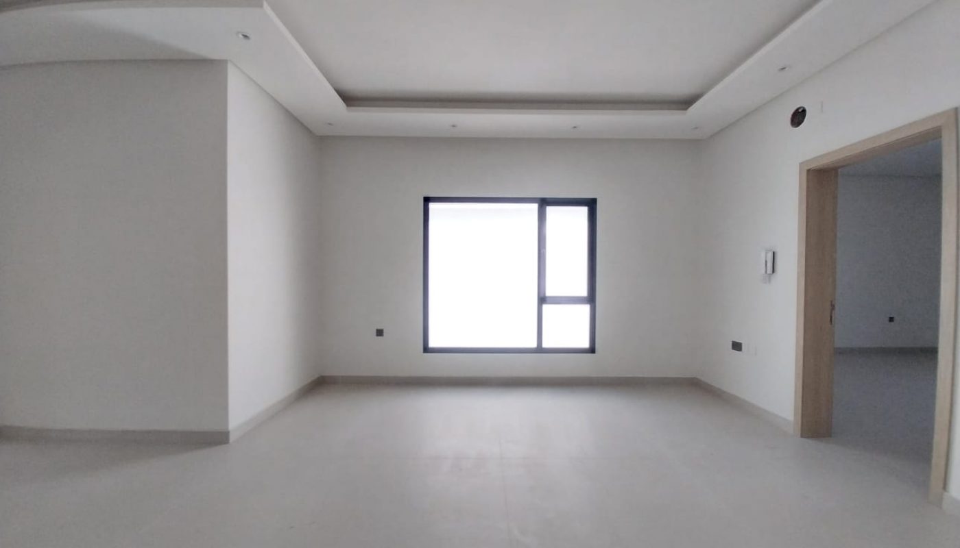 Empty, unfurnished room with light-colored walls and floor, a large window in the center, recessed ceiling lights, and a doorway on the right side.