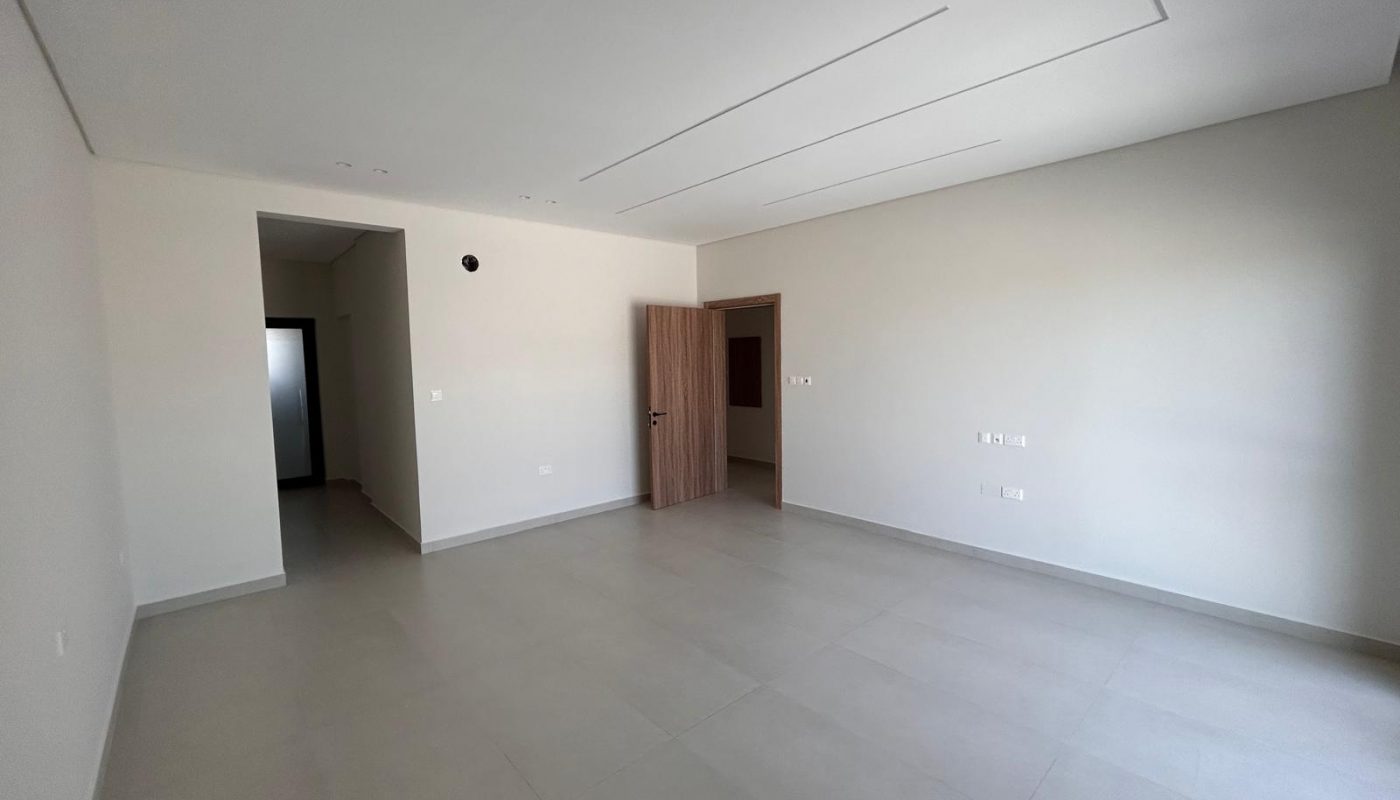 Empty, unfurnished room with light-colored tiled floor, white walls, an open wooden door, recessed ceiling, and visible electrical outlets.