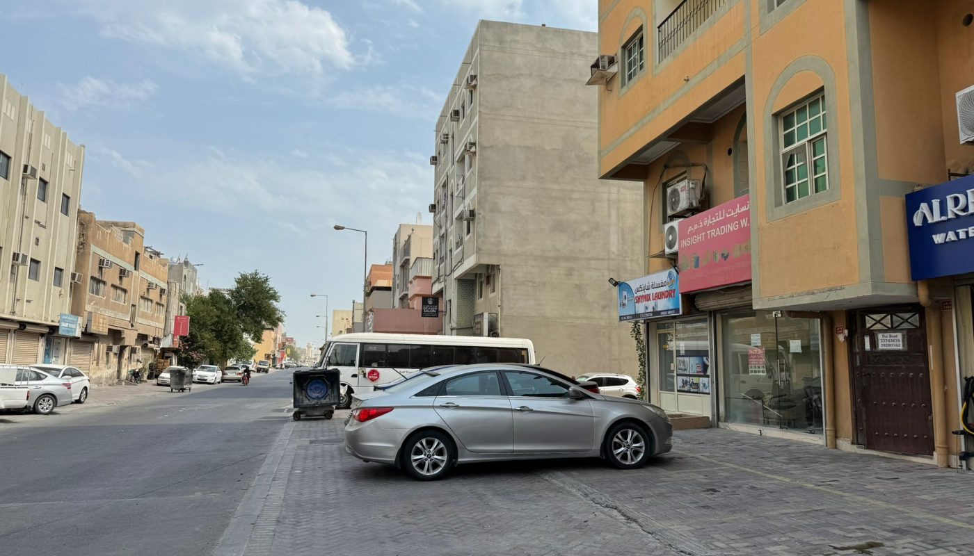 A silver car is parked on a sidewalk in front of a building with shops; a bus and garbage bin are visible on the street in the background.
