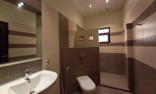 A modern bathroom with a white sink, a wall-mounted toilet, and a shower area separated by a tiled partition. The walls are tiled in brown and beige.